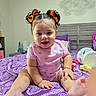 Cira Diaz is registered to the contest to win money with this photo: baby, child, girl, smiling, sitting, bed, bedroom, purple, hair_bows, onesie, patterned_bedspread, toy, furniture, indoor, cute, happy, person, infant, playful, decor