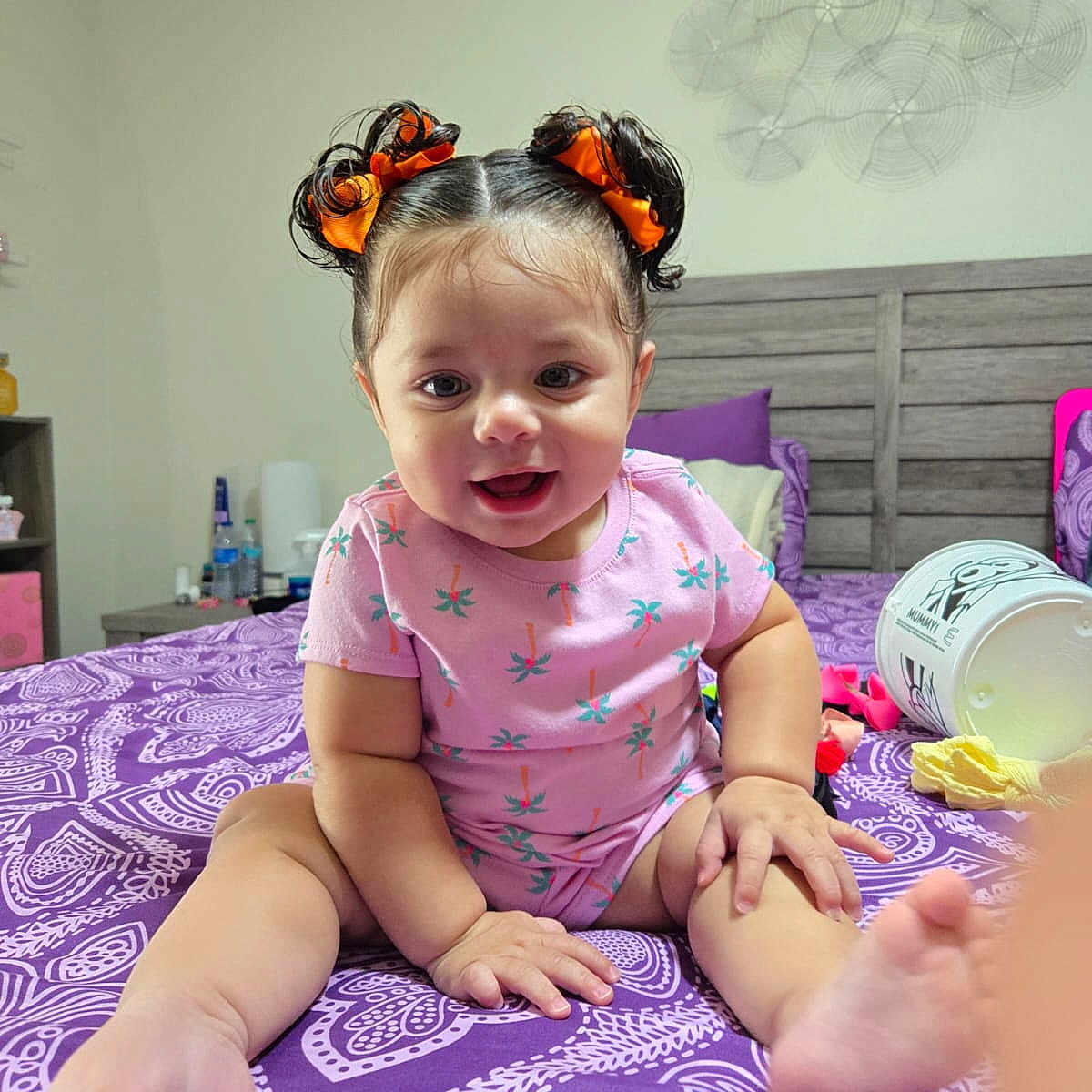 Cira Diaz is registered to the contest to win money with this photo: baby, child, girl, smiling, sitting, bed, bedroom, purple, hair_bows, onesie, patterned_bedspread, toy, furniture, indoor, cute, happy, person, infant, playful, decor