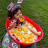 baby, infant, tub, orange_slices, strawberries, milk_bath, grass, headband, patriotic, lei, red_tub, outdoor, cute, child, summer, fruit, playful, celebration, milky_water, portrait