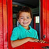 boy, bright_colors, casual_clothing, child, curly_hair, door, face, hand, happy, indoors, natural_light, playful, portrait, red_door, rustic, smiling, turquoise_shirt, window, wood, young