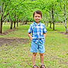 child, boy, smiling, standing, outdoor, greenery, trees, grass, plaid_shirt, shorts, sandals, nature, park, sunny, happy, portrait, casual_clothing, young, daylight, person