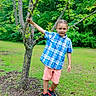 Memphis is registered to the contest to win money with this photo: child, boy, tree, greenery, grass, outdoor, smiling, plaid_shirt, shorts, sneakers, nature, park, leaves, young, standing, holding, casual_clothing, daylight, happy, person