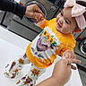 toddler, child, baby, laundromat, washing_machine, holding_hands, smiling, bow_headband, orange_clothing, sunflower_print, cow_print, standing, white_surface, adult_hand, indoor, floor, fabric, cute, happy, portrait