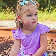 Miyah is registered to the contest to win money with this photo: child, girl, curly_hair, blue_bows, purple_shirt, striped_shorts, railroad_tracks, rocks, greenery, outdoor, sitting, portrait, daylight, nature, casual_clothing, young_child, expression, curious, summer, hair_ties