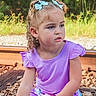 child, girl, curly_hair, blue_bows, purple_shirt, striped_shorts, railroad_tracks, rocks, greenery, outdoor, sitting, portrait, daylight, nature, casual_clothing, young_child, expression, curious, summer, hair_ties