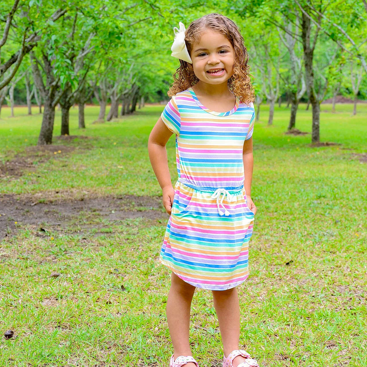 Miyah joined the competition — help win amazing prizes! child, girl, smile, curly_hair, bow, colorful_dress, sandals, grass, trees, orchard, outdoor, nature, greenery, sunlight, happy, standing, portrait, daylight, casual_clothing, young
