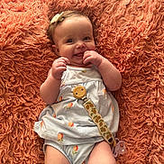 Ella is registered to the contest to win money with this photo: baby, infant, smile, pacifier, blanket, hair_bow, onesie, textured_background, cute, happy, portrait, lying_down, hands, skin, eyes, chubby_cheeks, peach_pattern, pacifier_clip, coral_blanket, cozy