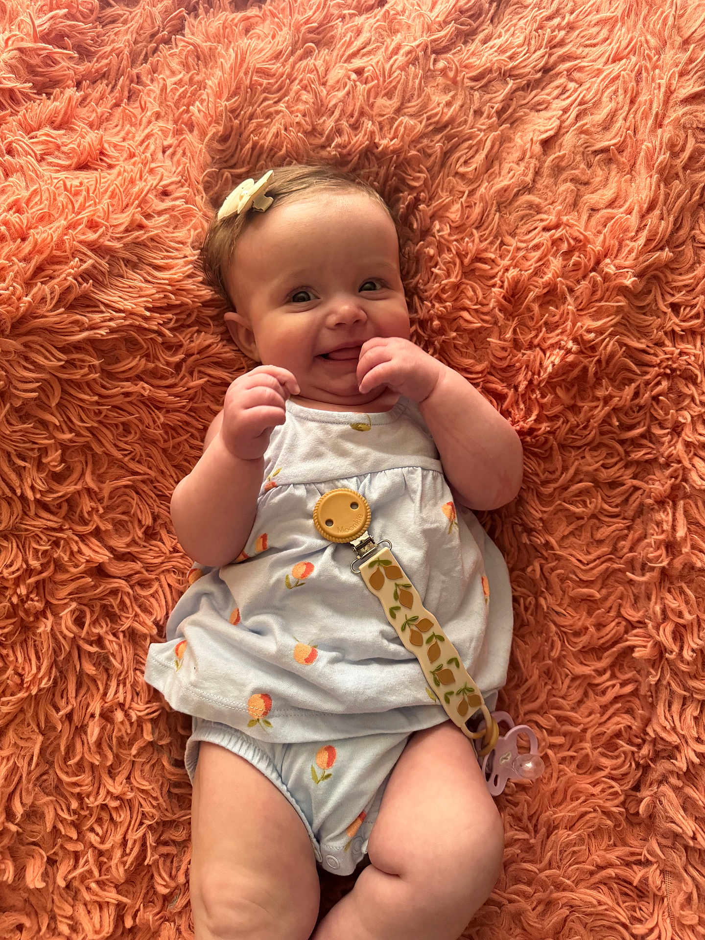 Ella is registered to the contest to win money with this photo: baby, infant, smile, pacifier, blanket, hair_bow, onesie, textured_background, cute, happy, portrait, lying_down, hands, skin, eyes, chubby_cheeks, peach_pattern, pacifier_clip, coral_blanket, cozy