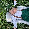 baby, blanket, blue_eyes, close_up, cute, flowers, grass, green_hat, green_pants, hat, infant, laying_down, leafy_ground, outdoors, portrait, shamrock, smile, spring, suspenders, white_shirt