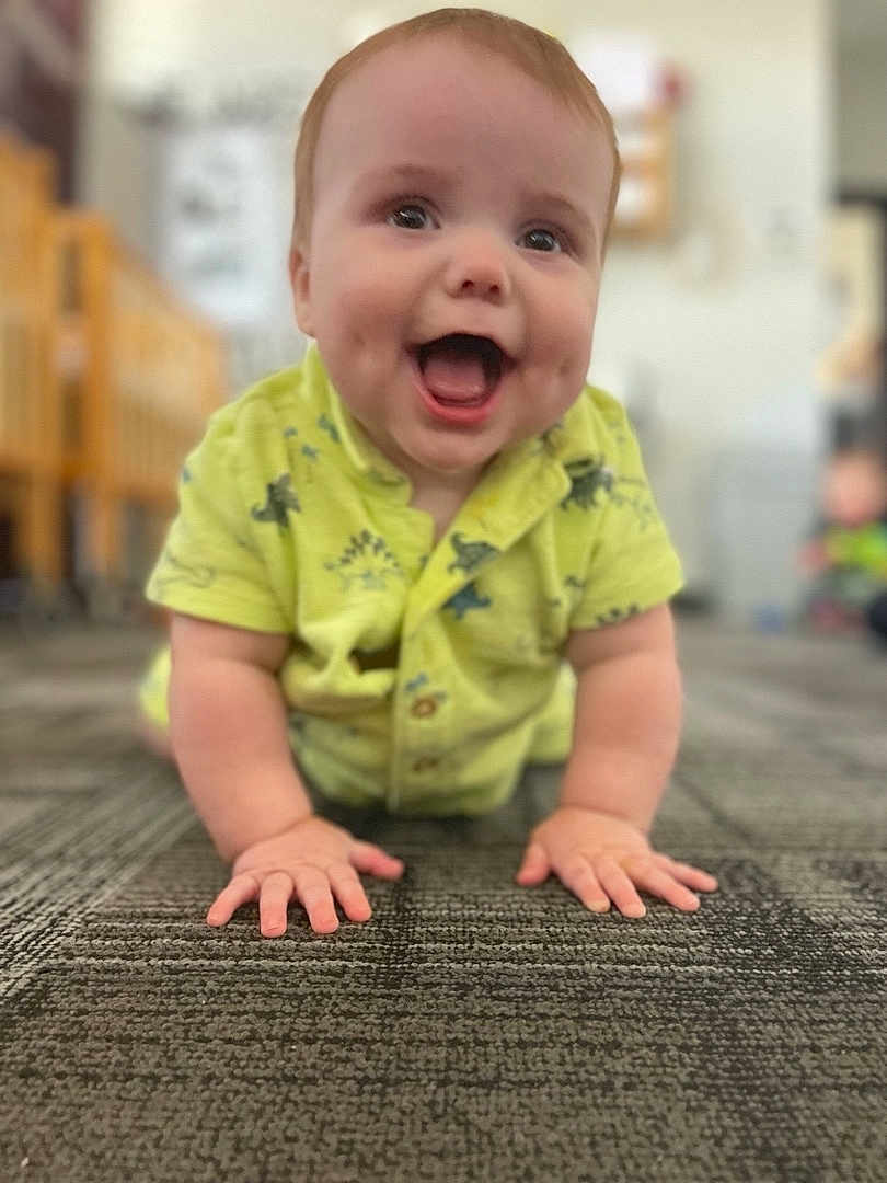 James is registered to the contest to win money with this photo: baby, child, crawling, smiling, happy, indoor, carpet, yellow_clothing, dinosaur_pattern, hands, face, cute, infant, young_child, portrait, closeup, playful, expression, person, toddler