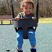 Andre is registered to the contest to win money with this photo: toddler, child, swing, playground, outdoor, smiling, happy, curly_hair, blue_jeans, black_shoes, black_swing, grass, bench, trees, sunny, daytime, person, park, casual_clothing, play