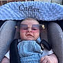 baby, child, car_seat, sunglasses, overalls, white_shirt, plush_headrest, straps, hand, face, brick_wall, outdoor, comfort, cute, infant, portrait, casual, fashion, seatbelt, blue