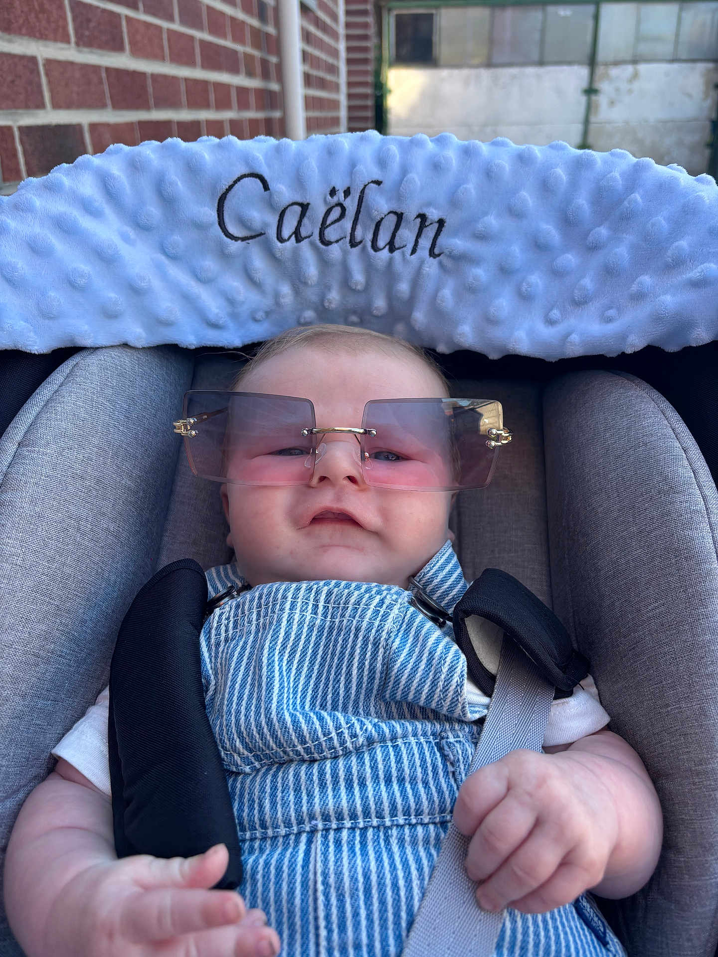 Caëlan a rejoint le concours — aidez-le/la à gagner de superbes lots ! baby, child, car_seat, sunglasses, overalls, white_shirt, plush_headrest, straps, hand, face, brick_wall, outdoor, comfort, cute, infant, portrait, casual, fashion, seatbelt, blue