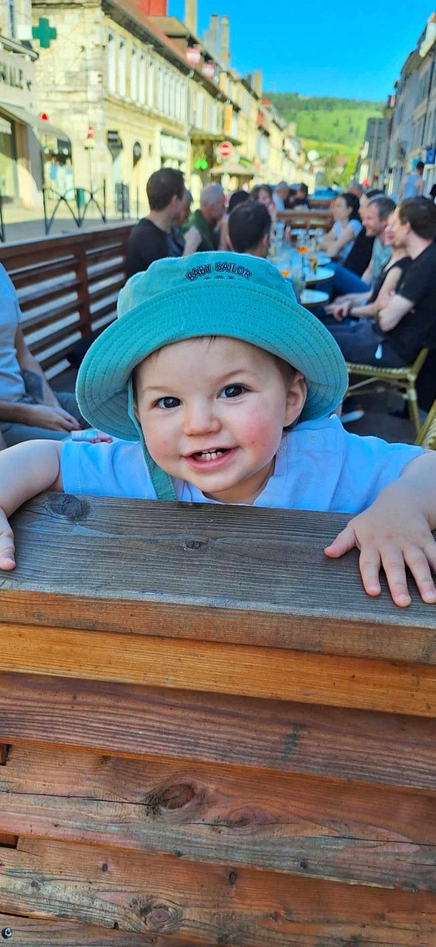 Léo a rejoint le concours — aidez-le/la à gagner de superbes lots ! toddler, child, hat, smile, outdoor, wood, fence, people, street, cafe, daylight, blue_sky, building, table, group, happy, casual, portrait, person, urban
