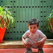 Aslan a rejoint le concours — aidez-le/la à gagner de superbes lots ! toddler, child, squatting, green_door, orange_pot, plant, indoor, curious, shoes, shorts, shirt, patterned_clothing, wide_eyes, red_floor, natural_light, decorative_door, hair, face, playful, casual