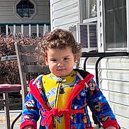 Elijah joined the competition — help win amazing prizes! toddler, child, curly_hair, robe, yellow_clothing, porch, outdoor, sunlight, chair, table, window, air_conditioner, railing, person, clothing, standing, daytime, expression, holding, background