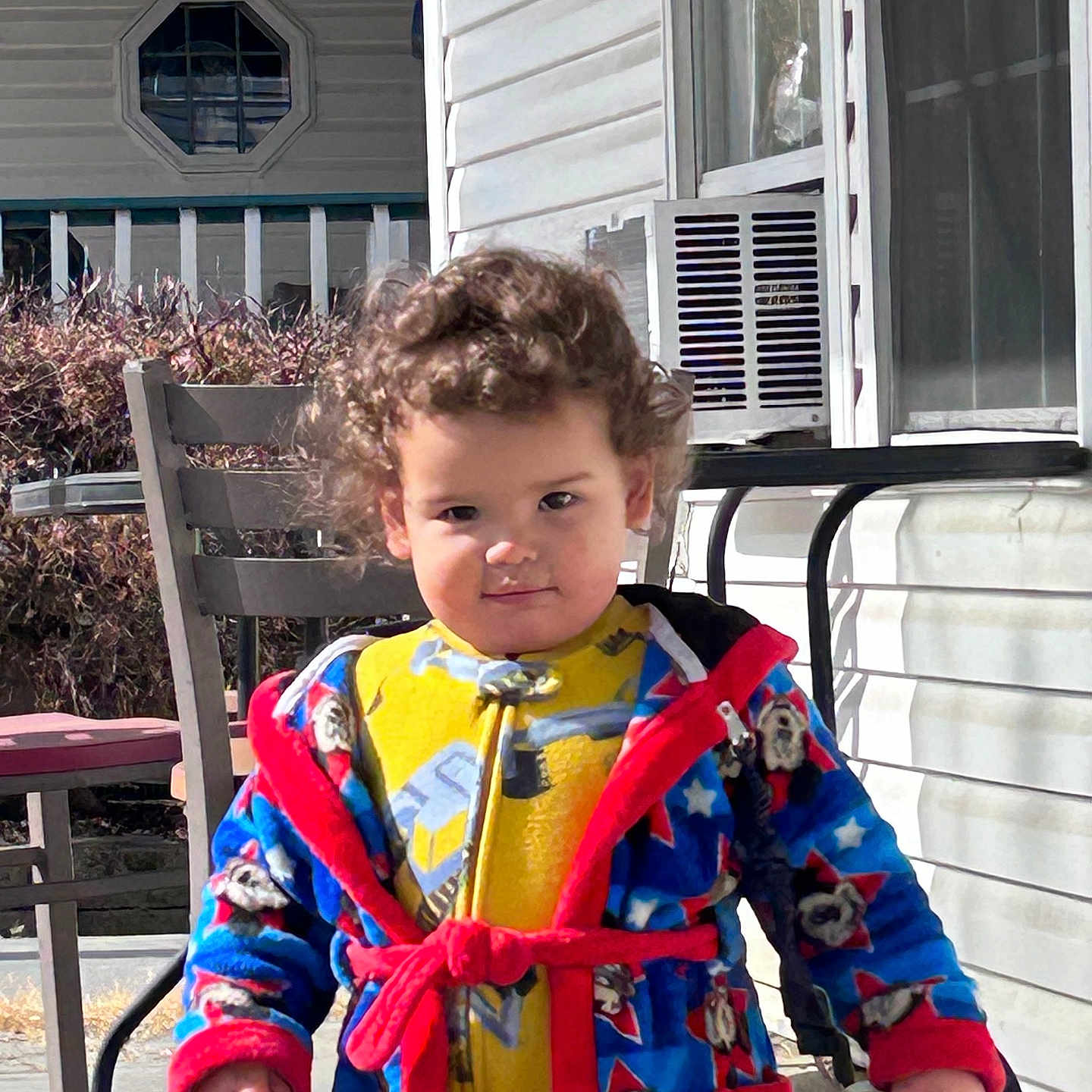 Elijah joined the competition — help win amazing prizes! toddler, child, curly_hair, robe, yellow_clothing, porch, outdoor, sunlight, chair, table, window, air_conditioner, railing, person, clothing, standing, daytime, expression, holding, background