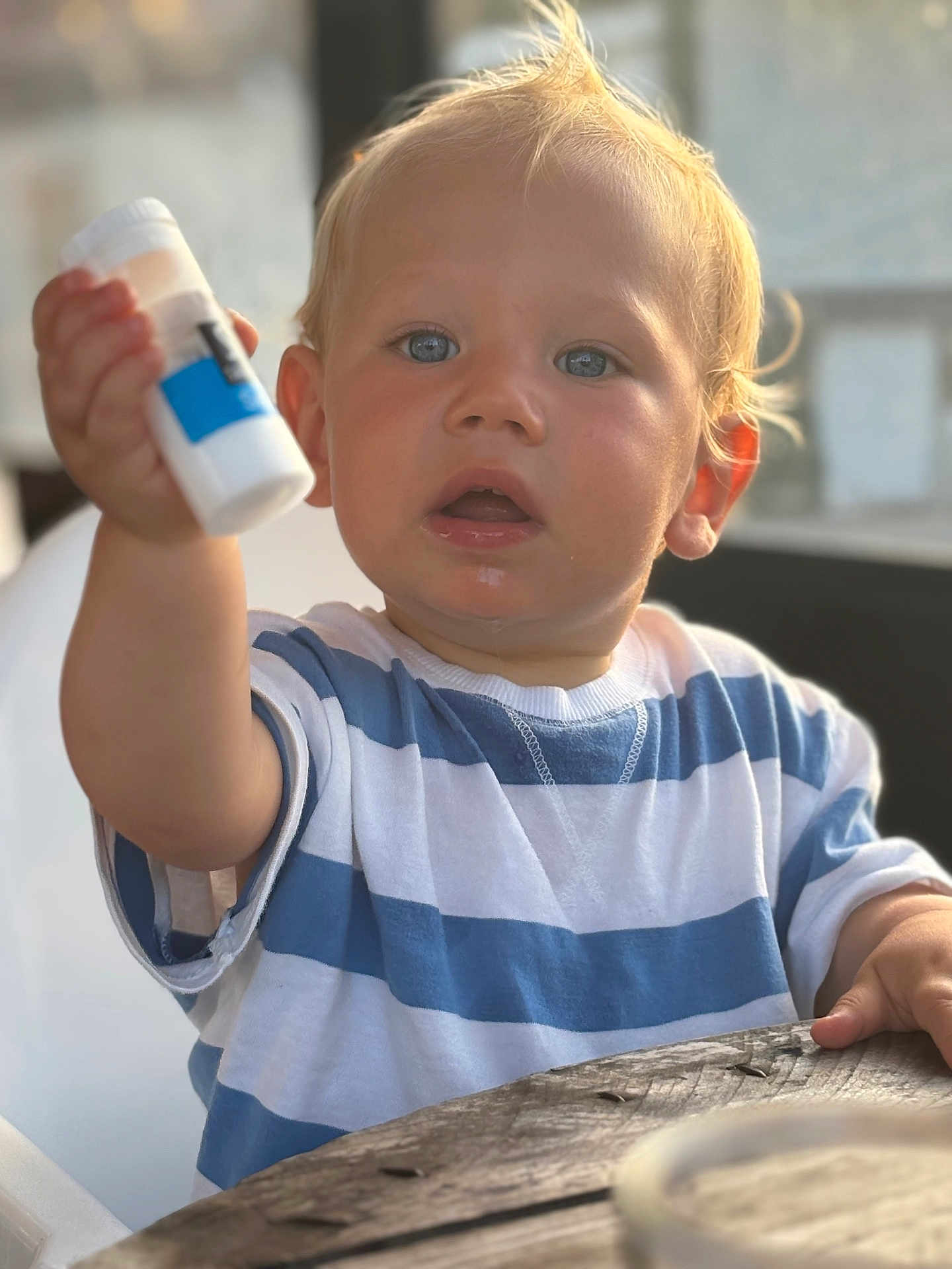 Alezio a rejoint le concours — aidez-le/la à gagner de superbes lots ! child, toddler, blue_eyes, blonde_hair, striped_shirt, white_and_blue, sitting, table, wooden_table, indoor, curious, holding_object, small_bottle, face, portrait, person, casual_clothing, young_child, expression, closeup