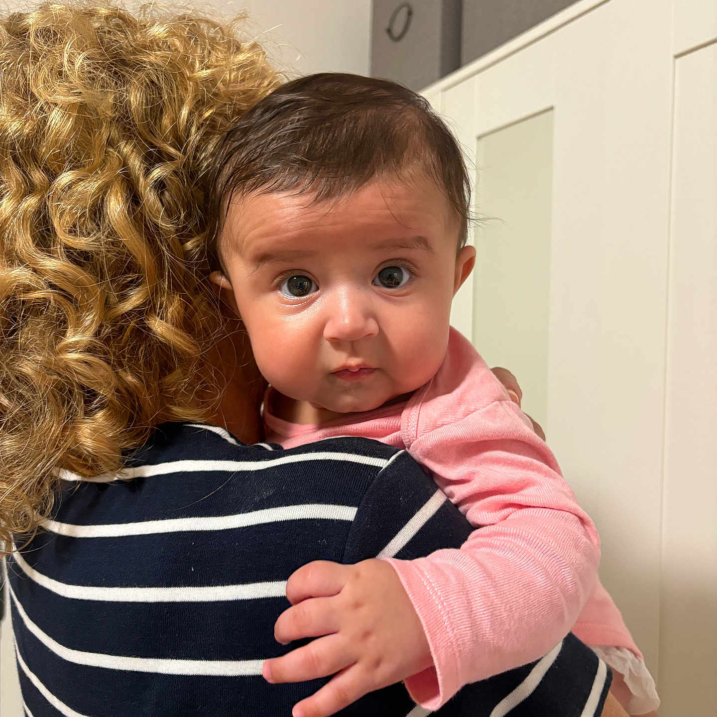 Mira Eliana is registered to the contest to win money with this photo: adult, baby, cabinets, child, close_up, curiosity, curly_hair, embrace, expression, face, hand, holding, home, indoor, person, pink_clothing, portrait, skin, striped_shirt, toddler