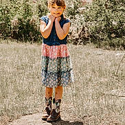 Khloey is registered to the contest to win money with this photo: child, girl, smiling, outdoor, field, grass, rocks, bushes, dress, colorful, cowboy_boots, sunny, nature, portrait, standing, happy, young, daylight, casual, cute