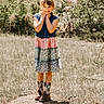 child, girl, smiling, outdoor, field, grass, rocks, bushes, dress, colorful, cowboy_boots, sunny, nature, portrait, standing, happy, young, daylight, casual, cute