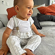 Siann participe au concours pour gagner de l'argent avec cette photo : toddler, child, smiling, sitting, striped_pants, suspenders, white_shirt, couch, pillows, living_room, happy, barefoot, indoor, modern_interior, face, person, home, cute, casual_clothing, relaxed