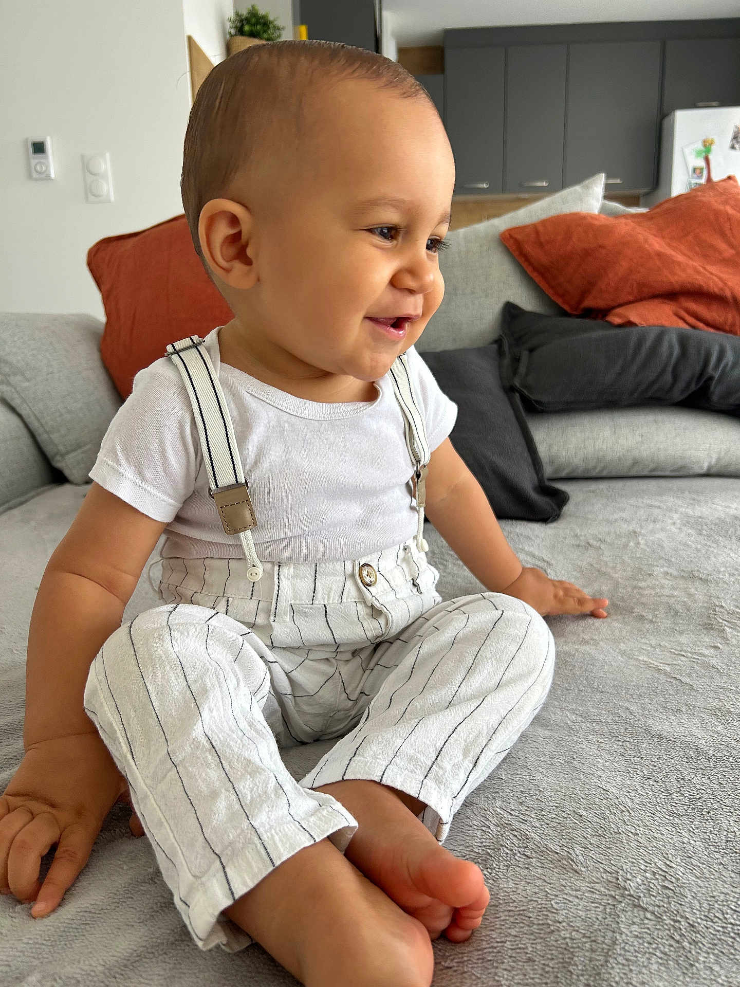 Siann participe au concours pour gagner de l'argent avec cette photo : toddler, child, smiling, sitting, striped_pants, suspenders, white_shirt, couch, pillows, living_room, happy, barefoot, indoor, modern_interior, face, person, home, cute, casual_clothing, relaxed