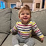 baby, child, smile, happy, sitting, couch, living_room, sweater, striped_clothing, gray_pants, white_socks, furniture, indoor, person, hair, face, expression, cozy, home, cute