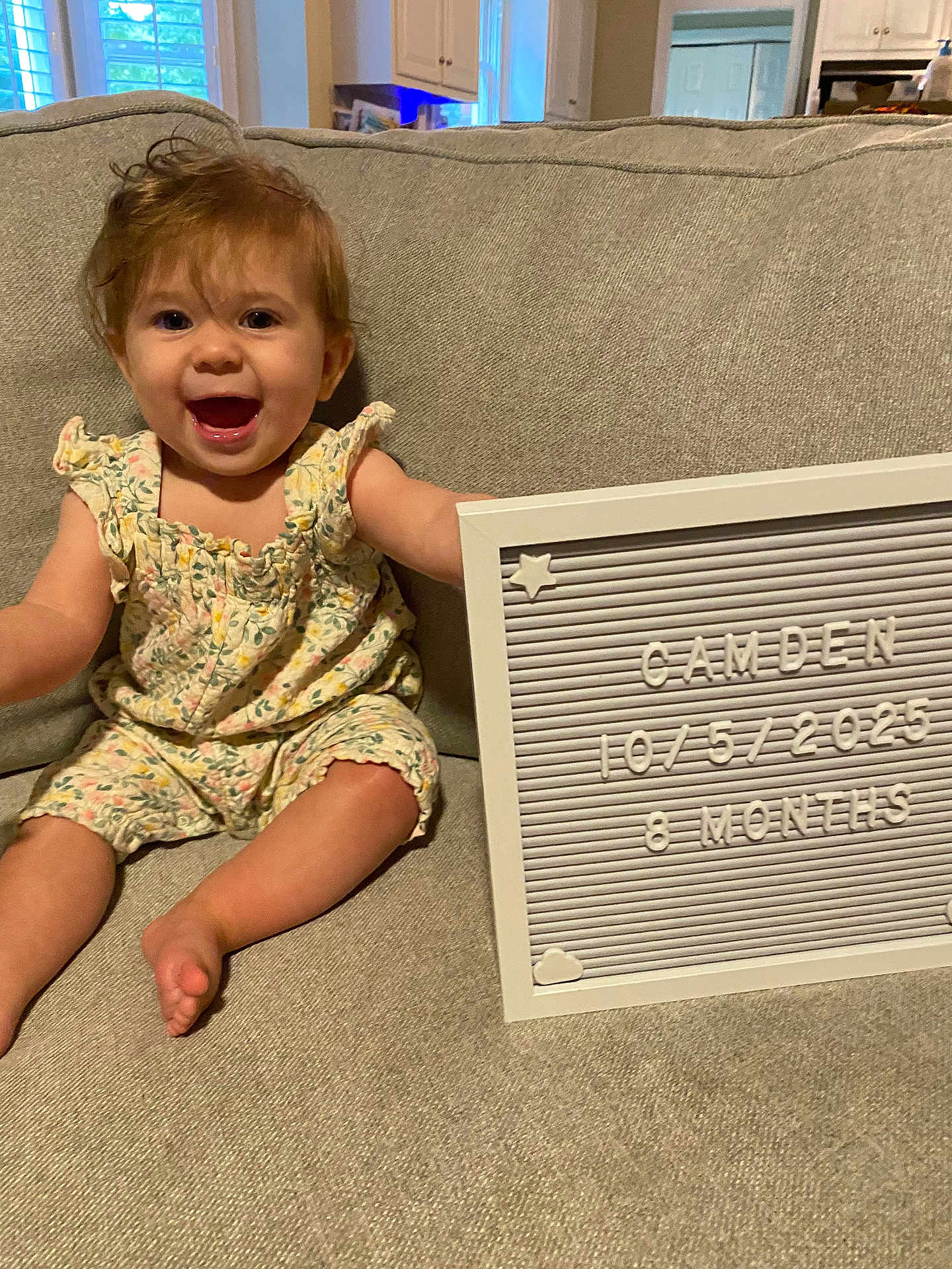 Camden is registered to the contest to win money with this photo: baby, child, smiling, couch, romper, letter_board, indoor, happy, face, person, living_room, hair, clothing, foot, sitting, milestone, home, furniture, cute, infant