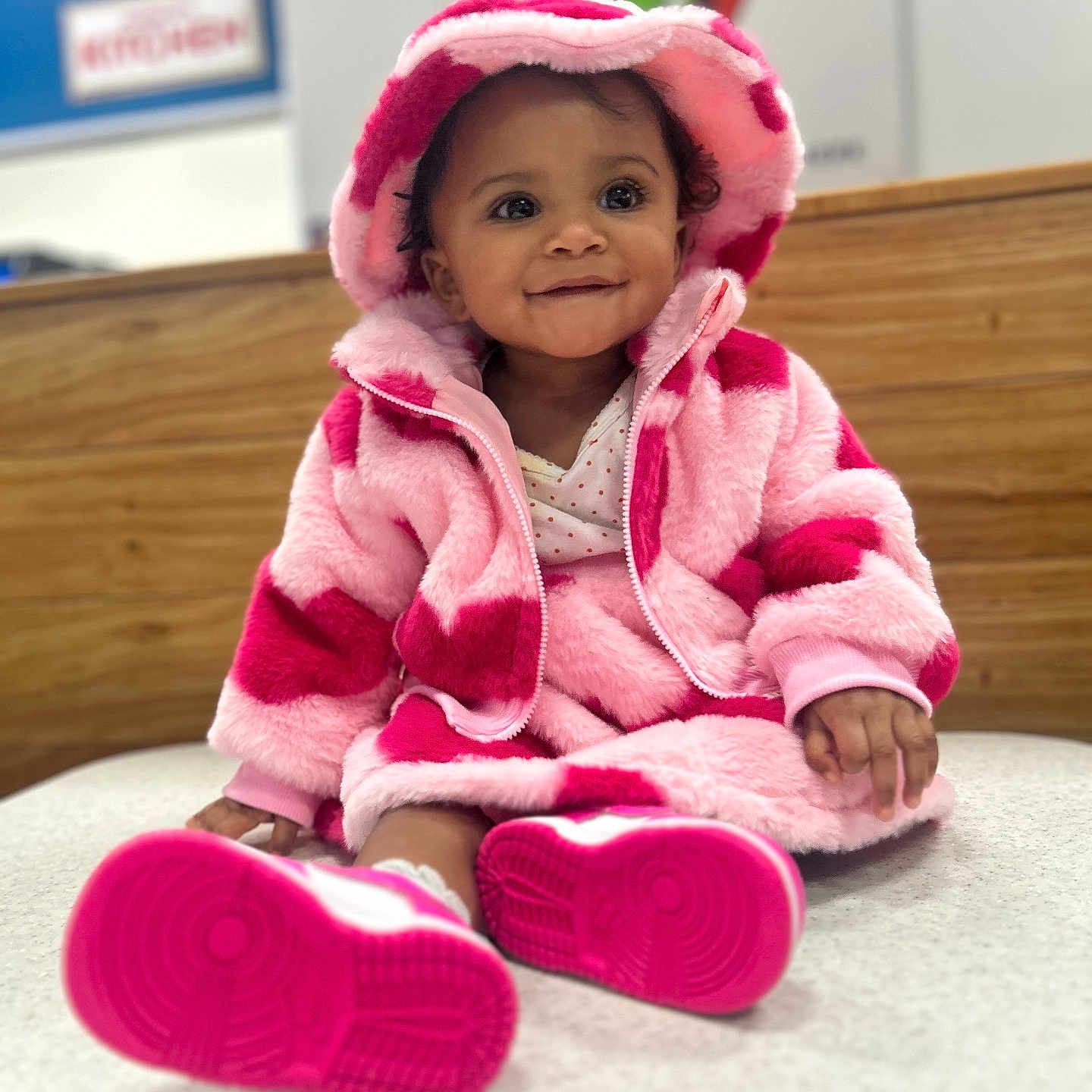 Makani is registered to the contest to win money with this photo: baby, bonnet, clothing, coat, face, footwear, hardwood, hat, head, hood, hoodie, indoors, knitwear, person, photography, portrait, shoe, sweater, sweatshirt, wood