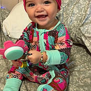Kamaiyah is registered to the contest to win money with this photo: baby, child, smile, pajamas, headwrap, toy, bed, blanket, happy, sitting, indoor, face, person, colorful_clothing, cute, infant, bracelet, foot, patterned_fabric, home