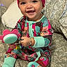 baby, child, smile, pajamas, headwrap, toy, bed, blanket, happy, sitting, indoor, face, person, colorful_clothing, cute, infant, bracelet, foot, patterned_fabric, home