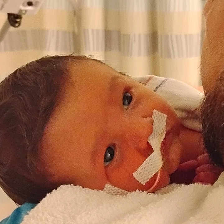 Elijah is registered to the contest to win money with this photo: adult, baby, blanket, care, close_up, curtains, eyes, face, healthcare, holding, hospital, indoor, infant, medical_tape, nasal_tube, newborn, person, rest, tender_moment, wrap