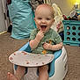 baby, child, smiling, sitting, baby_seat, snack, food, green_shirt, carpet, indoor, lamp, storage_bin, laundry, clutter, happy, cute, infant, feeding, playful, toddler