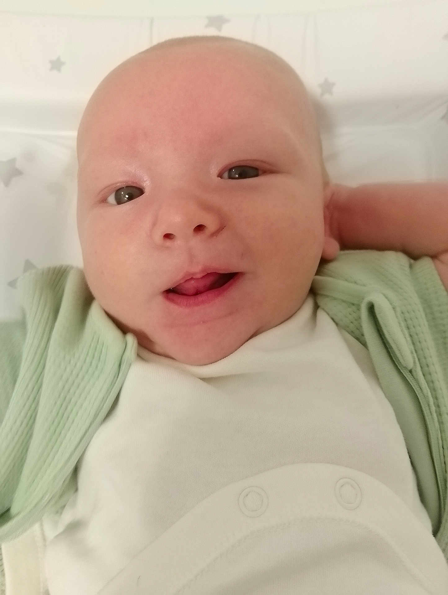 Luca is registered to the contest to win money with this photo: baby, infant, face, smiling, clothing, white_outfit, green_cardigan, close_up, skin, head, eyes, mouth, lying_down, star_pattern, background, cute, child, newborn, person, portrait
