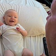 Luca is registered to the contest to win money with this photo: adult, baby, bed, chair, clothing, face, folded_clothes, furniture, hand_holding, happy, indoor, infant, interaction, onesie, person, pillow, remote_control, skin, smile, tattoo
