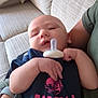 baby, pacifier, arm, couch, pillow, clothing, person, sleepy, indoor, fabric, hand, face, infant, sitting, relaxing, comfort, casual, adult, wristwatch, text