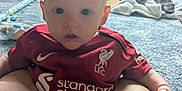 Luca is registered to the contest to win money with this photo: baby, baby_swing, casual, child, curtains, floor, home, indoor, infant, legs, liverpool_jersey, living_room, looking_at_camera, person, red_shirt, rug, seated, slippers, toy, window