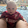 baby, baby_swing, casual, child, curtains, floor, home, indoor, infant, legs, liverpool_jersey, living_room, looking_at_camera, person, red_shirt, rug, seated, slippers, toy, window