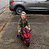 car, casual_clothing, child, daytime, happy, hood, hooded_jacket, outdoor, pavement, person, play, sidewalk, small_bike, smiling, street, toddler, toy_motorcycle, transportation, urban, vehicle