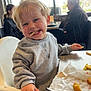 toddler, child, high_chair, food, french_fries, fried_food, restaurant, indoor, casual, messy_face, smiling, blond_hair, sweatshirt, table, window, lamp, people, seated, daylight, hand