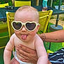 baby, sunglasses, heart_shaped, tongue_out, diaper, tattoo, adult_hand, outdoor, greenery, table, chair, baby_bottle, summer, funny_face, cute, child, person, playful, sunlight, casual