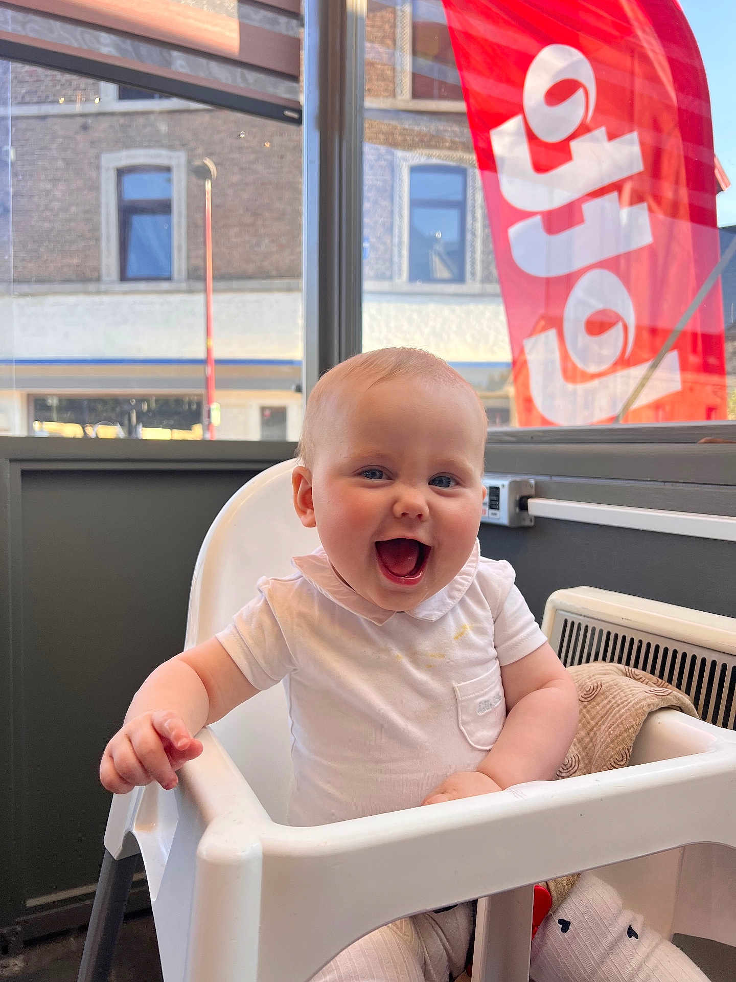 Lexie participe au concours pour gagner de l'argent avec cette photo : baby, high_chair, smiling, indoor, window, red_flag, white_shirt, child, happy, face, person, furniture, building, light, seat, clothing, tongue, skin, hair, background