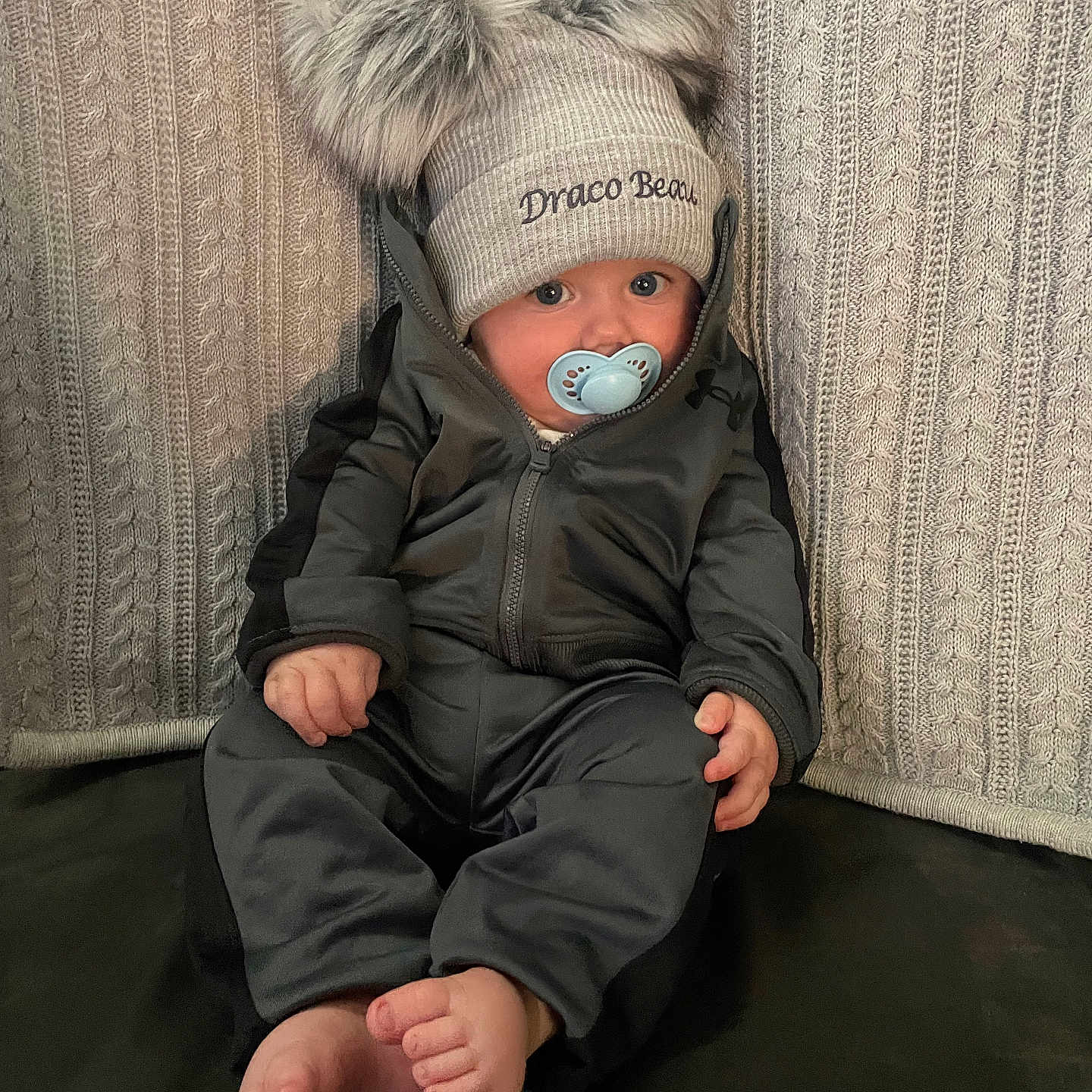 Draco is registered to the contest to win money with this photo: baby, barefoot, blanket, child, curious, cute, face, feet, hands, hat, indoor, jacket, knit, pacifier, pom_poms, portrait, sitting, texture, warm_clothing, young