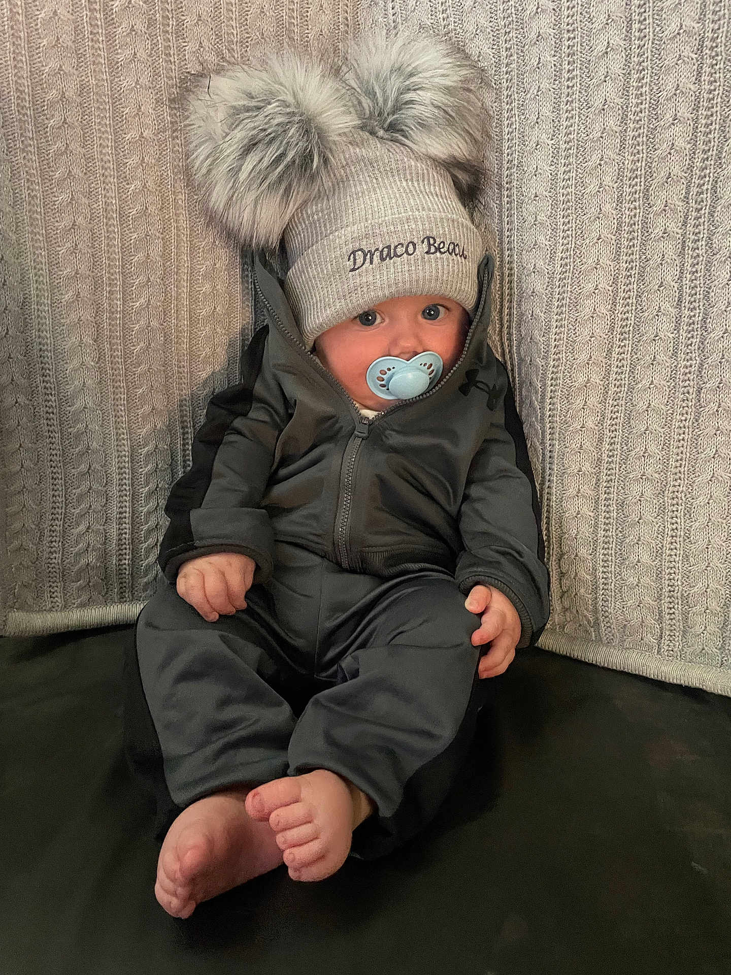 Draco is registered to the contest to win money with this photo: baby, pacifier, hat, pom_poms, knit, jacket, child, sitting, barefoot, blanket, texture, indoor, cute, curious, portrait, warm_clothing, face, hands, feet, young