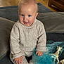 baby, child, knitted_clothing, blue_cloth, giraffe_toy, sitting, indoor, soft_surface, happy, smiling, cozy, cute, toddler, floor, wood_floor, cushion, sock, hand, face, portrait