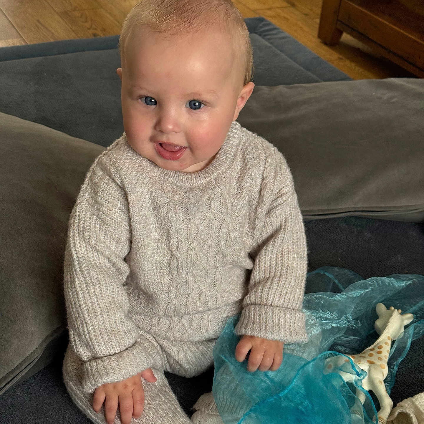 Jackson is registered to the contest to win money with this photo: baby, blue_cloth, child, cozy, cushion, cute, face, floor, giraffe_toy, hand, happy, indoor, knitted_clothing, portrait, sitting, smiling, sock, soft_surface, toddler, wood_floor