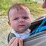 baby, infant, face, expression, outdoor, nature, grass, gravel, person, holding, wrap_carrier, clothing, hand, skin, head, blue_eyes, furrowed_brow, adult, closeup, sunlight
