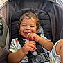 toddler, child, smile, curly_hair, car_seat, seatbelt, snack, happy, cute, infant, clothing, baby, hands, face, sitting, indoor, young_child, cozy, playful, joy