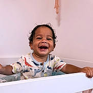 Takoah is registered to the contest to win money with this photo: toddler, child, smiling, laughing, crib, baby, indoor, clothing, dinosaur_pattern, happy, portrait, face, hand, young_child, room, furniture, cute, curly_hair, skin, buttoned_shirt